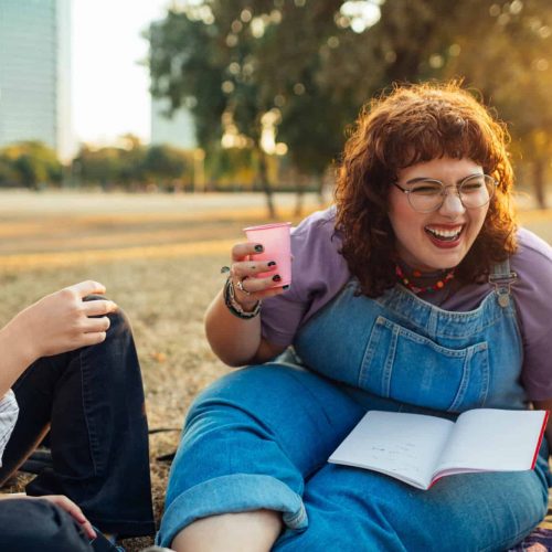 Students sitting in park, studying together. Portrait of beautiful plus size girl holding plastic cup and laughing