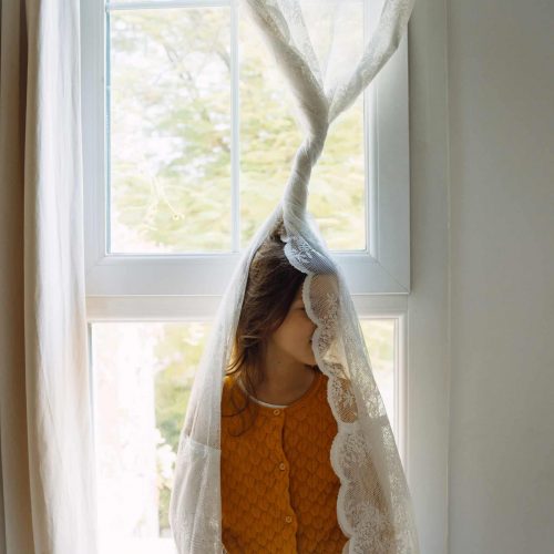 A horizontal portrait of a little girl hiding behind the curtain lace in the room
